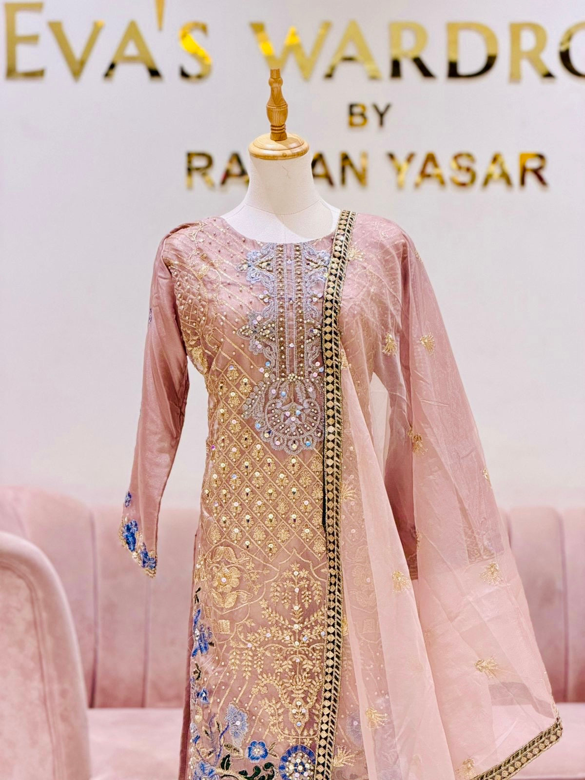 Blush pink embroidered women’s ethnic suit displayed on a mannequin, featuring intricate gold threadwork, mirror detailing, floral motifs, and a sheer matching dupatta with a decorative border, showcased inside Eva’s Wardrobe 